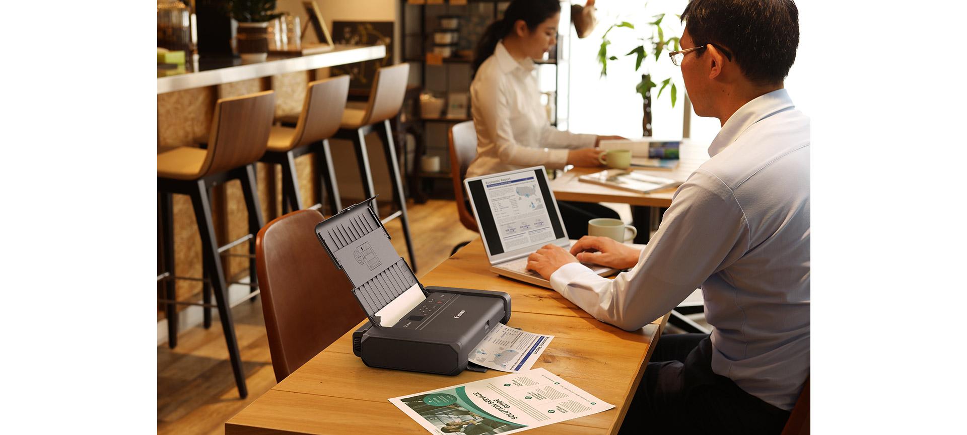 Homme assis dans un café travaillant sur son ordinateur portable et imprimant un fichier avec une imprimante portable sans fil Canon PIXMA TR150.