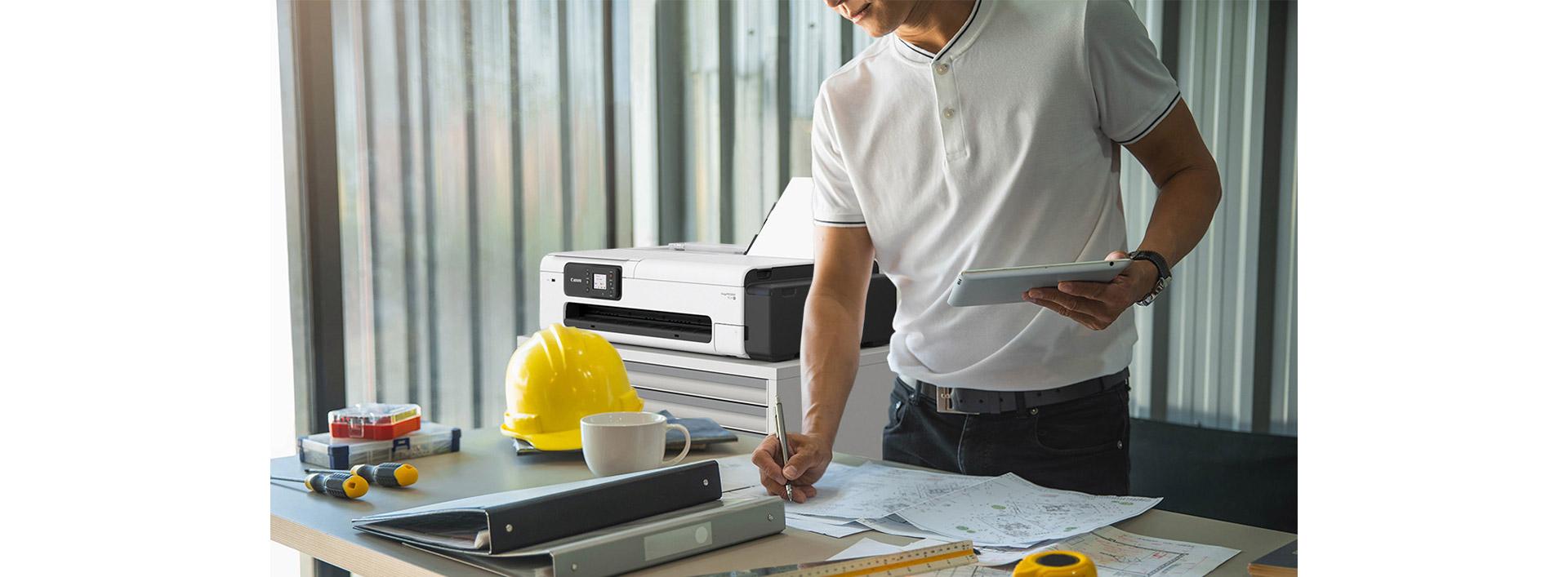 Architecte debout derrière son bureau en train de dessiner des plans. L’imprimante grand format Canon imagePROGRAF TC-21 est posée sur une étagère derrière lui.