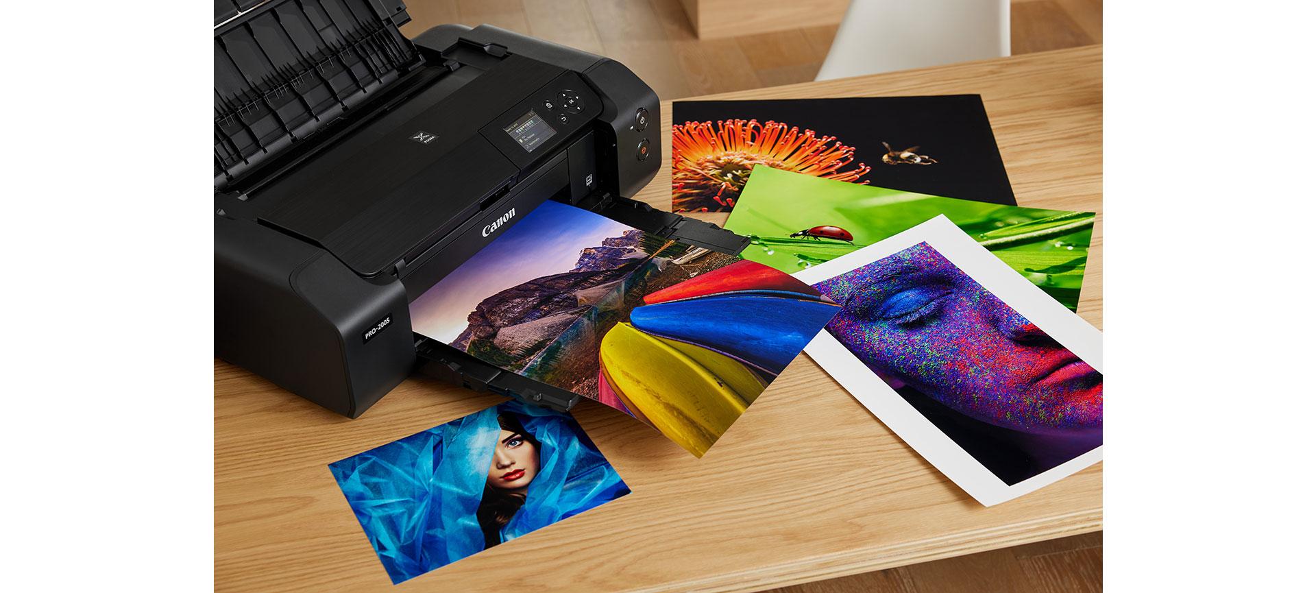 A Canon imagePROGRAF PRO-200S photo printer outputs a vibrant mountain photo on a wooden desk alongside several colourful art prints.