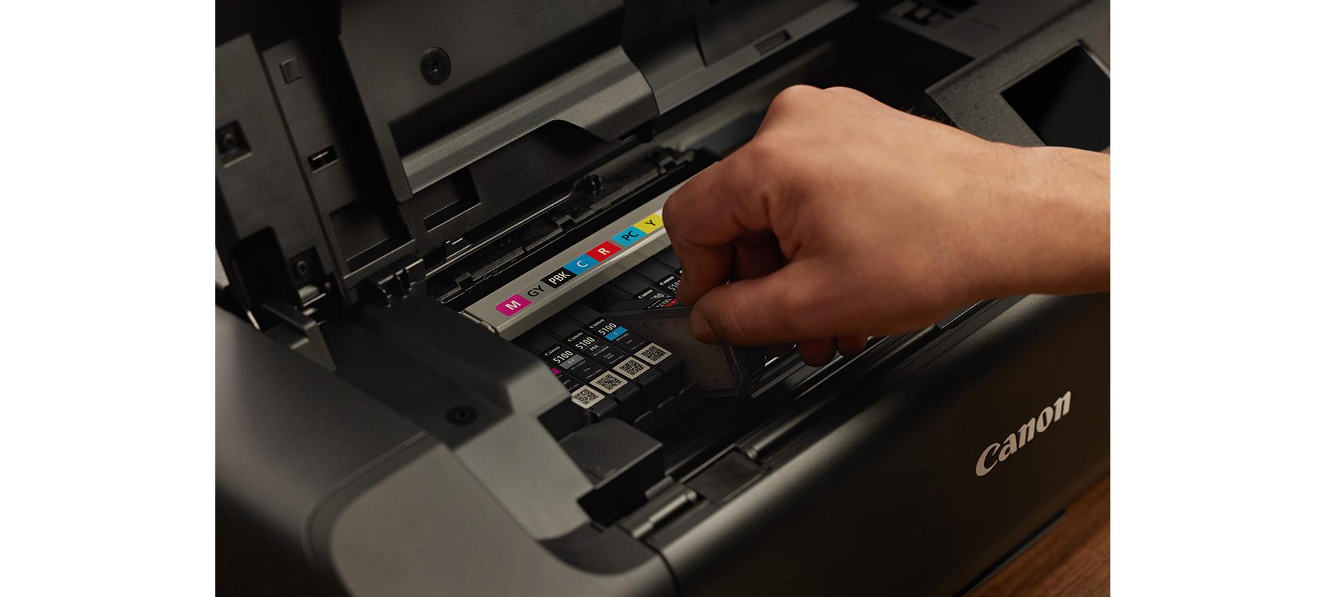 A hand installs an ink cartridge into the open colour compartment of a Canon photo printer.