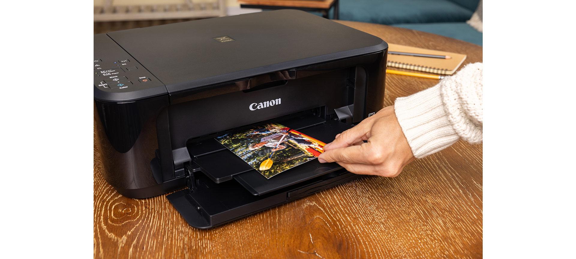 A hand retrieves a freshly printed colour photo of a kayaker from the tray of a black Canon PIXMA printer.